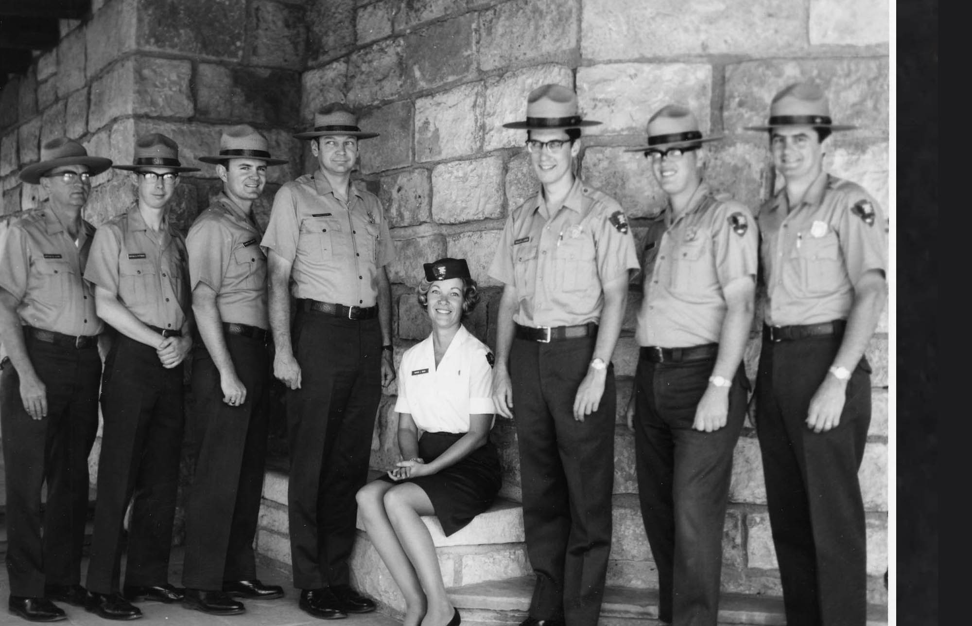 Marion Riggs in NPS uniform skirt, blouse, small arrowhead pin, and pillbox hat sits between seven men standing in uniforms with shield-shaped badges.